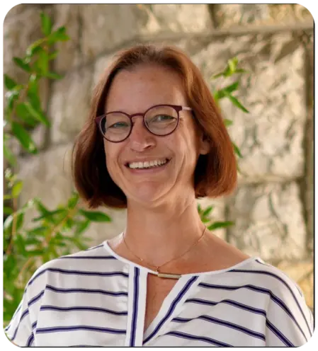 Porträt von Susan Zeiter, Administration und Kasse beim Verein Autismus Kunterbunt. Sie hat rötlich-braunes Haar, trägt eine Brille und ein gestreiftes Oberteil und lächelt freundlich vor einem Hintergrund aus Naturstein und Pflanzen.